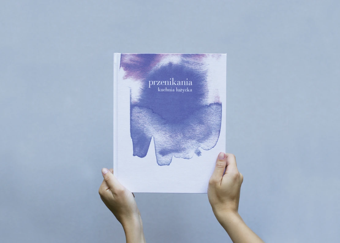 Hands holding the book „przenikania – kuchnia łużycka”, its cover washed in blue watercolour, against a grey wall