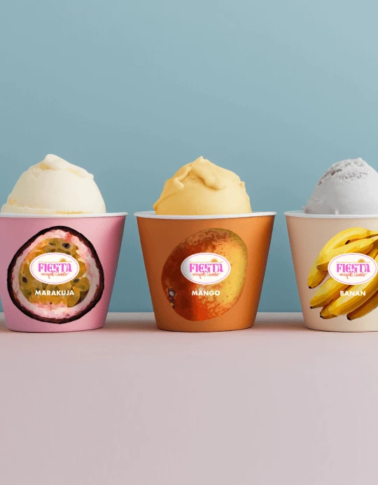 Three Fiesta ice cream cups in a row on a blue background — marakuja, mango and banan — each cup wearing its fruit illustration